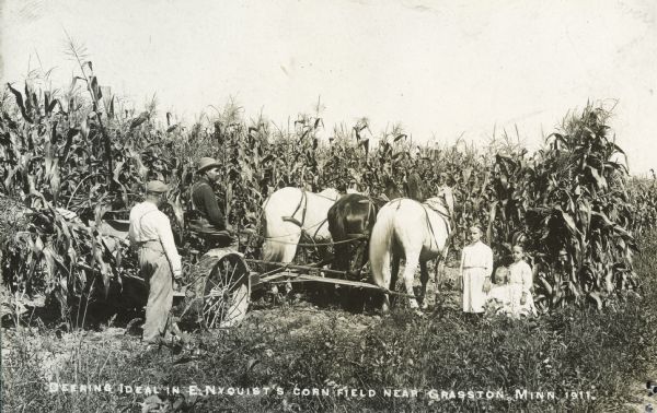 A man and three children gather around a man operating a horse-drawn Deering Ideal corn binder in E. Nyquist's cornfield.