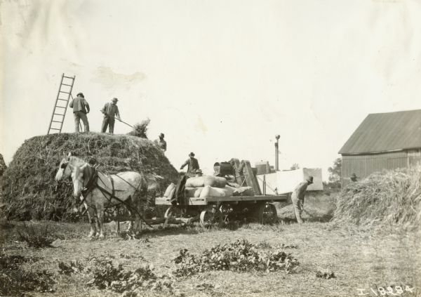 A group of farmers feeds grain into a thresher with a horse-drawn wagon nearby.