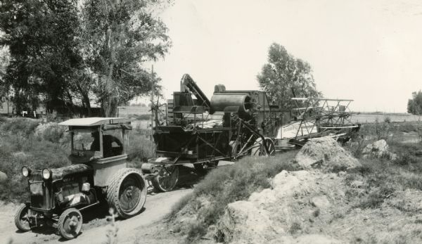 Three-quarter view from front left of a man using a tractor to pull a McCormick-Deering harvester-thresher (combine) down a dirt road.