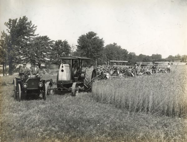 A tractor pulls a series of four grain binders while a man drives an IHC Auto Wagon alongside and a crowd of men looks on.