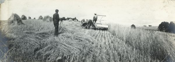 A man photographs a grain binder at work in a field.