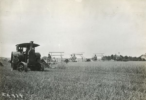 Three grain binders follow a Mogul Junior tractor.  A farmhouse among trees is in the background.
