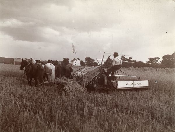 An American flag flies from a McCormick grain binder as it is pulled through a field by four horses.