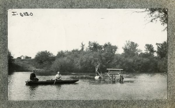 A man leads two horses pulling a rice(?) binder through water while two others in a nearby canoe look on.