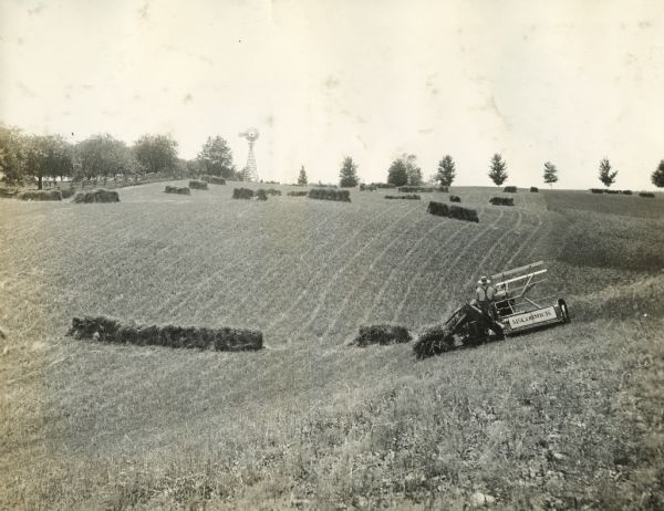 A farmer uses a McCormick grain binder on a hillside with a windmill in the background.