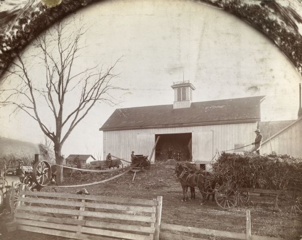 Farmers using a husker-shredder(?) powered by an engine. A man and two children look on from the doorway of a barn. Another man is standing on a horse-drawn wagon.