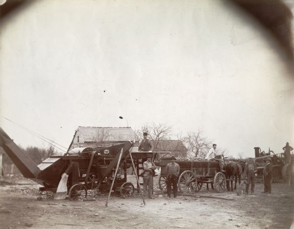 A group of men and a dog gather around a husker-shredder and wagon.