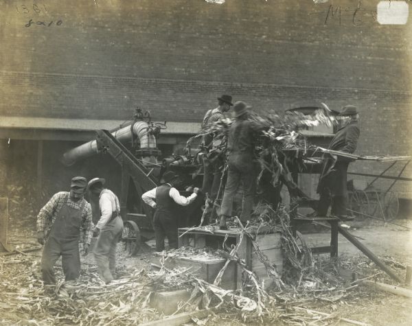 A group of men loading corn husks into a husker-shredder near a barn.