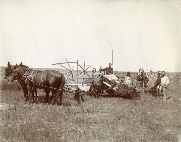 Four men and a woman gather around a horse-drawn McCormick grain binder in a field. Two men are sitting in a horse-drawn wagon.