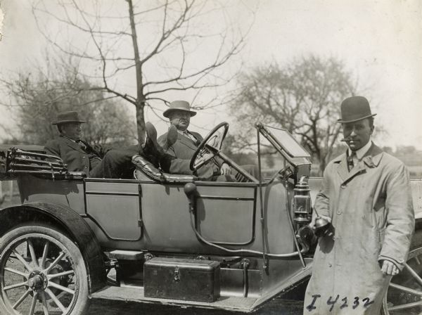 Two men relax in an automobile while another stands by with an oil can in hand, smoking a cigar.