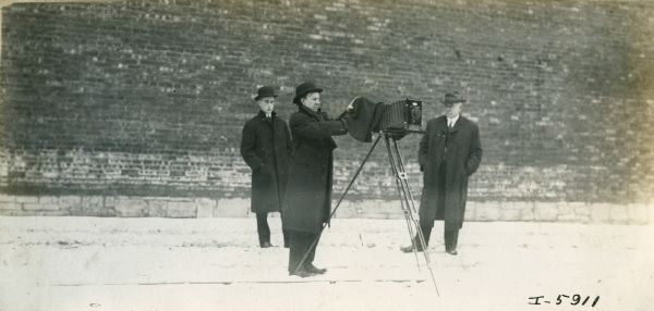 A man prepares a camera to take a photograph, while two other men stand behind him. The man is likely an International Harvester company photographer. The location is likely McCormick Works in Chicago.