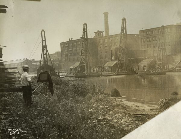 A photographer is standing near a camera on the bank of the Chicago River. He likely works for International Harvester Company and appears to be photographing the company's McCormick Works on the opposite shore. A sign on one of the buildings reads: "Driver No. 5 Great Lakes Dredge & Dock Co."
