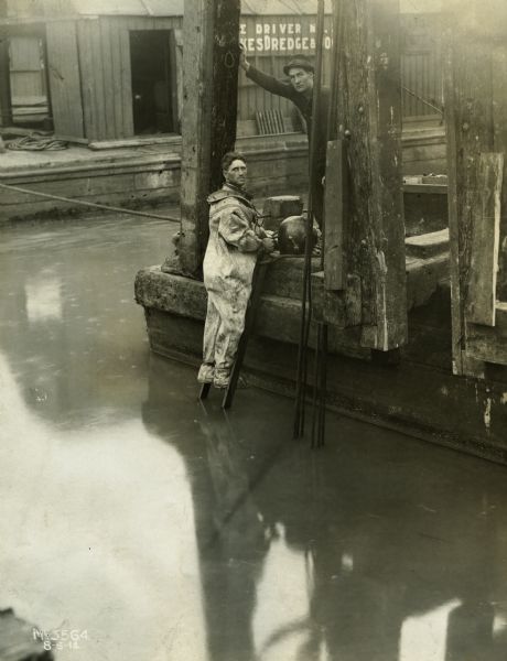 A man wearing a diving suit stands on a ladder coming out of a body of water (possibly the Chicago River). A sign in the background reads, "Dredge". The man likely worked for the Great Lakes Dredge and Dock Company.
