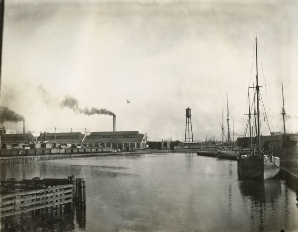 Chicago River near International Harvester's McCormick Works (factory). A sailboat, water tower, railroad cars and many industrial buildings line the shore.