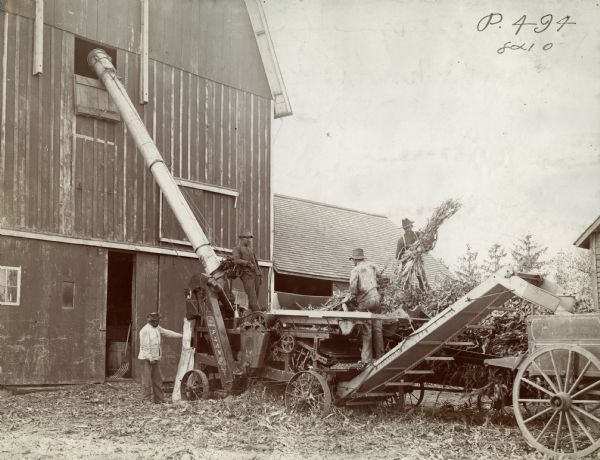 Farmers operating a Plano husker-shredder near a barn.
