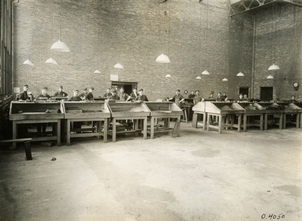 Workers in the core room of the "malleable shop" at Osborne Works. Large lamps with white shades are hanging from the tall ceiling over the work tables.