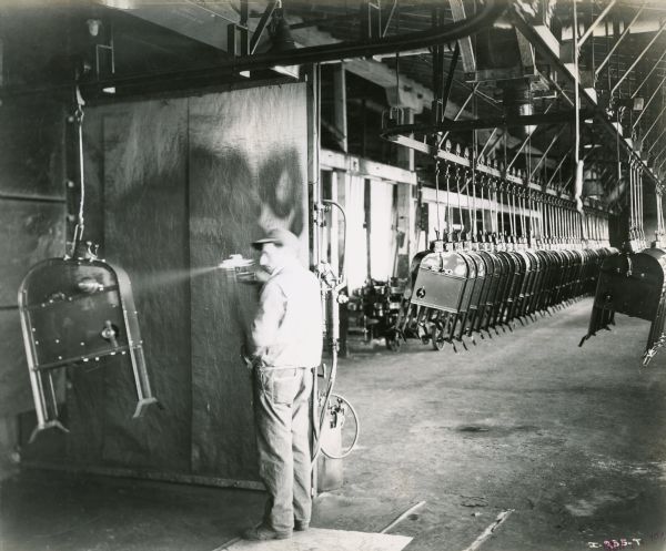 Factory worker spray painting a corn sheller at International Harvester's Rock Falls Works. Corn shellers are suspended from the ceiling in the background.