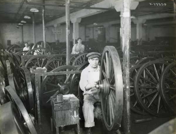 Man working on a wagon wheel at International Harvester's Weber Works. Two other workers and a fire escape sign are in the background.