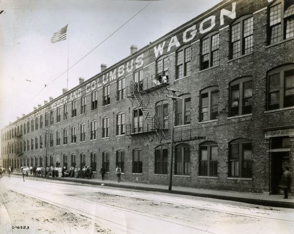 Men line up outside on the sidewalk in front of the International Harvester's Weber Works building. The factory produced wagons. Two men are looking down from windows on the third floor which has a fire escape. An American flag is flying from a flagpole on the roof.