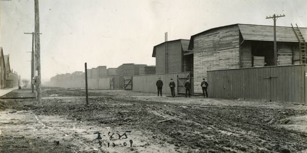 View across unpaved road towards four men standing outside of the Weber Works lumberyard. The men are standing near a gate in the fence surrounding industrial buildings. The factory produced wagons.
