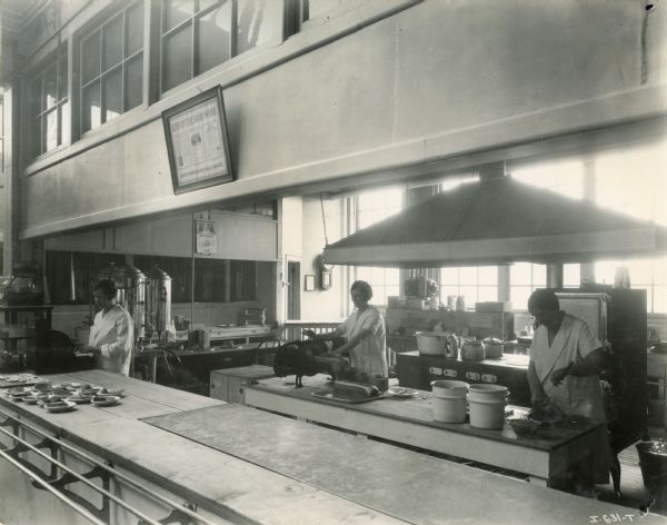 Three women work behind the counter of the Hamilton Works (factory) cafeteria. A sign hanging on the wall reads, "Keep up the good work.  Design, workmanship, sales, service." The kitchen calendar advertises, "Duff's - ever since 1867."