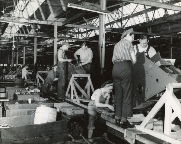 Male and female factory workers assemble corn pickers at Chatham Works. According to the original caption, the work was "lightened" by a roller-conveyor assembly line, electric hoists, and electric nut-setters.