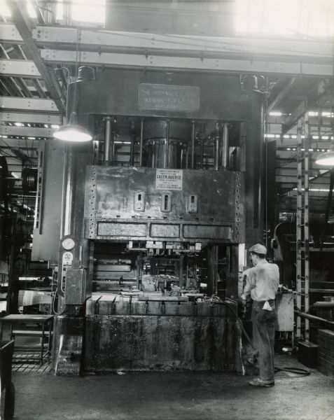Factory worker operating a 500 ton triple-action hydraulic press at Chatham Works. According to the original caption, the second pass has completed the forming of folder sheet-metal canvas-coupler of a harvester-thresher (combine). The final shape is such that the completed part must be pushed off the die by the air cylindar at left of the photograph. The operator is protected by a photo-electric light curtain.