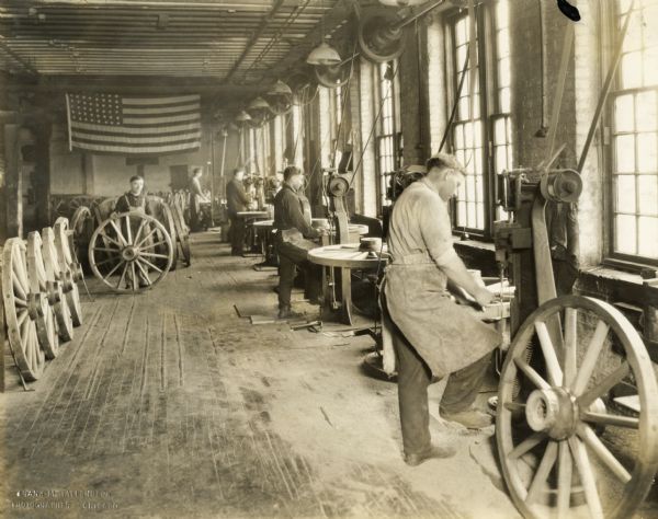 Factory workers making wagon wheels at International Harvester's Weber Wagon Works. The men are working near the windows. An American flag is hanging on the wall in the background.