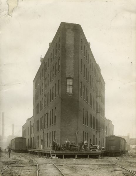 Exterior view of an International Harvester Transfer Agency building with several men standing on a platform surrounding it. Railroad tracks are located on either side of the building.