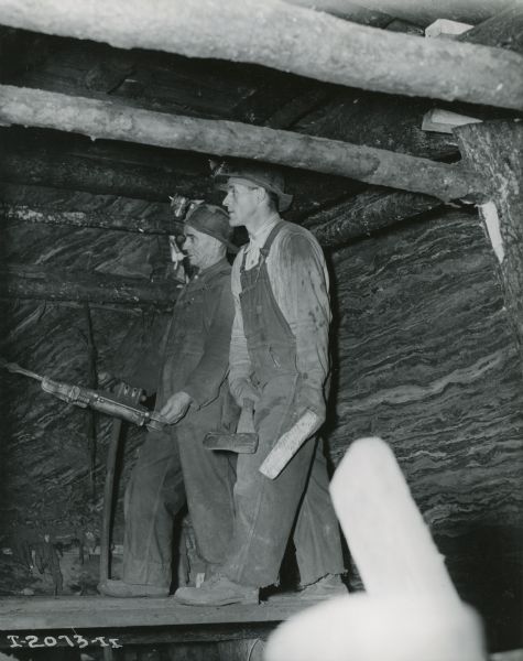Two workers, both in overalls and hardhats, use tools underground at International Harvester's Hawkins Mine in Hibbing, Minnesota. One man uses a drill while the other holds an ax.