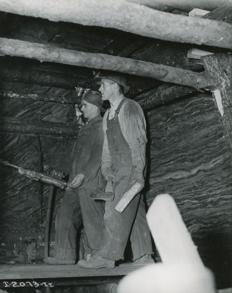 Two workers, both in overalls and hardhats, use tools underground at International Harvester's Hawkins Mine in Hibbing, Minnesota.  One man uses a drill while the other holds an ax.