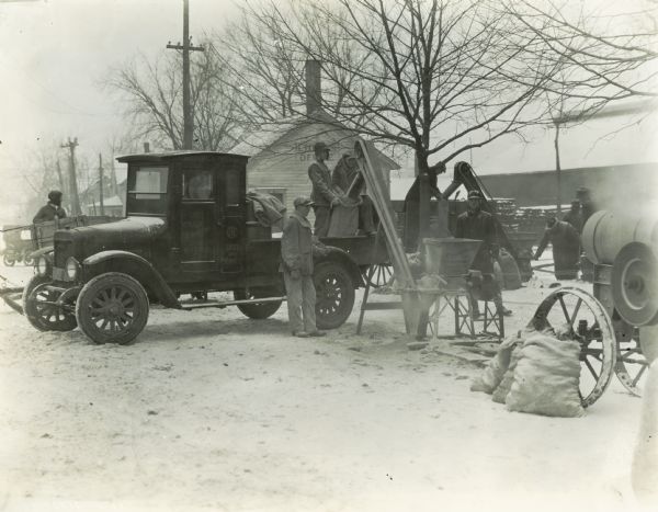 Group of men using an International feed grinder outside a lumber office, possibly in Iowa. Some of the men are standing on top of a truck owned by Milo E. Smith Sales and Service, an International Harvester dealership.