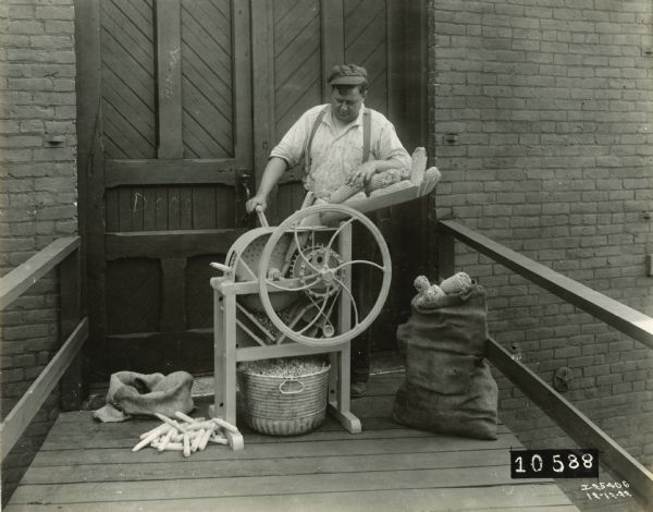 Man operating a Keystone corn sheller manufactured by International Harvester. The man is standing in front of a brick building.