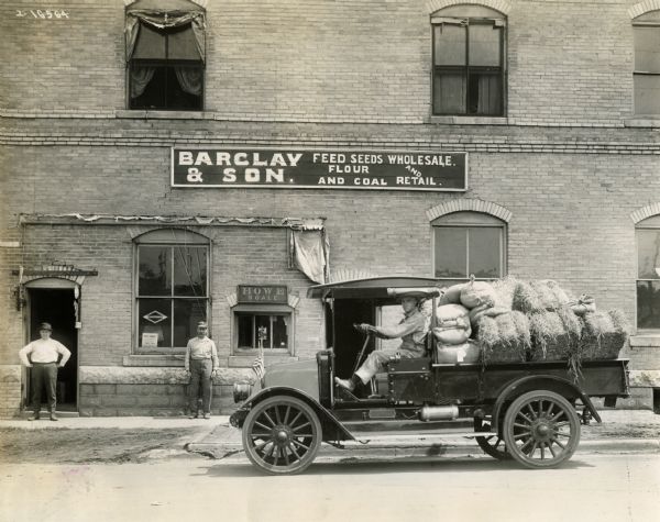 Two men standing on the sidewalk outside of the Barclay & Son Wholesale and Retail store. Another man is sitting in what is possibly an International H-21 truck loaded with bales of hay and grain.