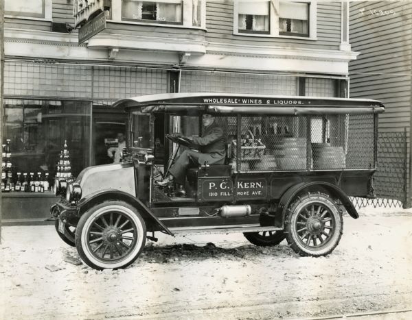 A man is sitting in the driver's seat of an International truck owned by P.C. Kern. The truck is filled with barrels and bottles of wines and liquors and is parked on a snow-covered street. The truck is parked in front of the P.C. Kern Wholesale Wines & Liquors store at 1310 Fillmore Avenue.