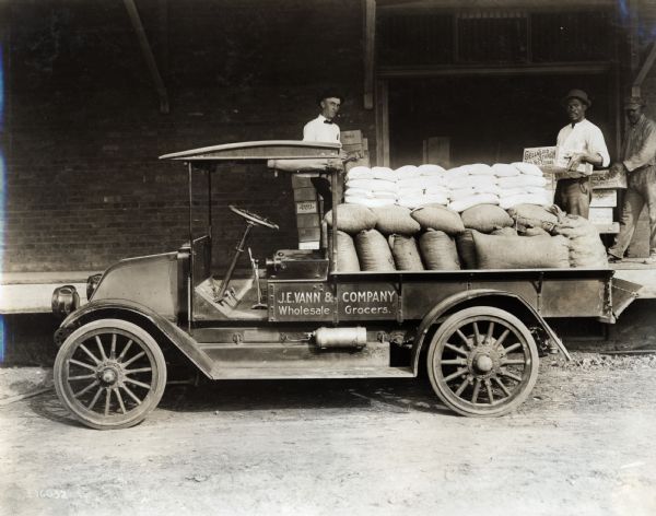 Three men unload an International truck owned by J.E. Vann and Company Wholesale Grocers. The truck is carrying bags and crates of merchandise and is parked in a store loading dock, presumably in Vero Beach.
