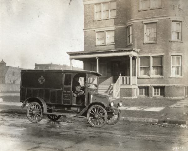 A man sits in an International truck outside of a large three-story house in Chicago. The truck bears the text "The Fair."