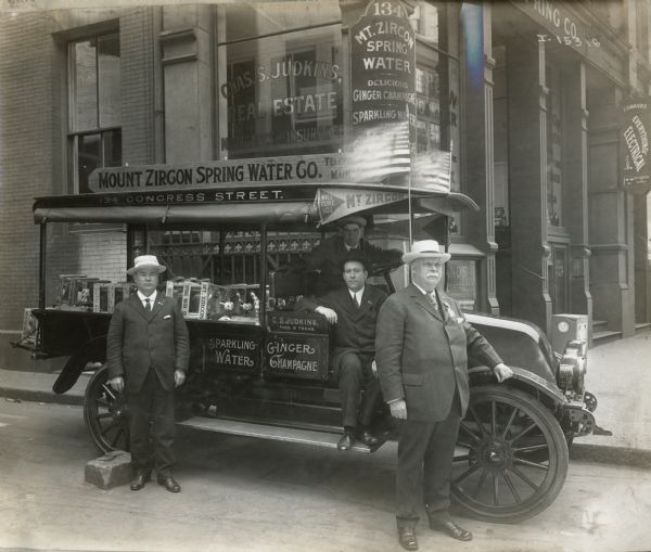 Four men pose around an International truck, probably a Model H, owned and operated by the Mount Zircon Spring Water Co. The truck is loaded with cases and bottles of sparkling water and ginger champagne. Behind them is the company's corner store, Chas. S. Judkin's Real Estate office, and Edmand's Everything Electrical shop. The older man in the photo may be Charles S. Judkins.