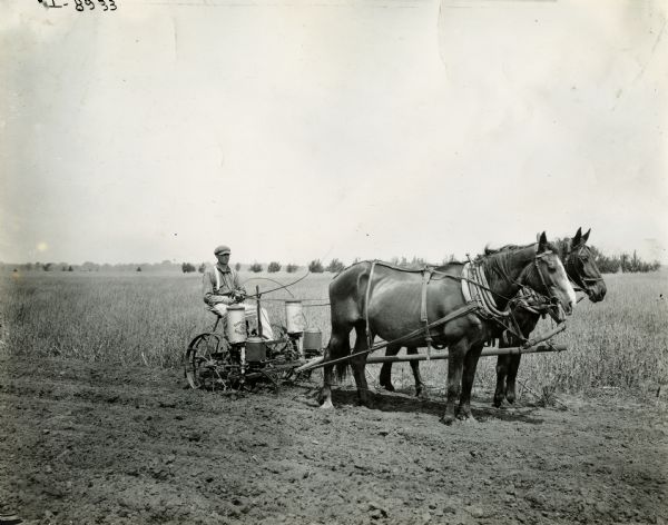 A man sitting on an International Harvester corn planter pulled by two large horses.