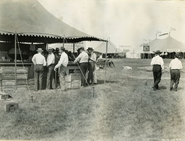 A group of men stand at a makeshift bar under a beverage tent where crates from Golden Grain Juice Company and Anheuser-Busch from St. Louis, Missouri, line the ground. The tent may be part of a agricultural exhibition or fair. A tent in the background bears the name "Avery."