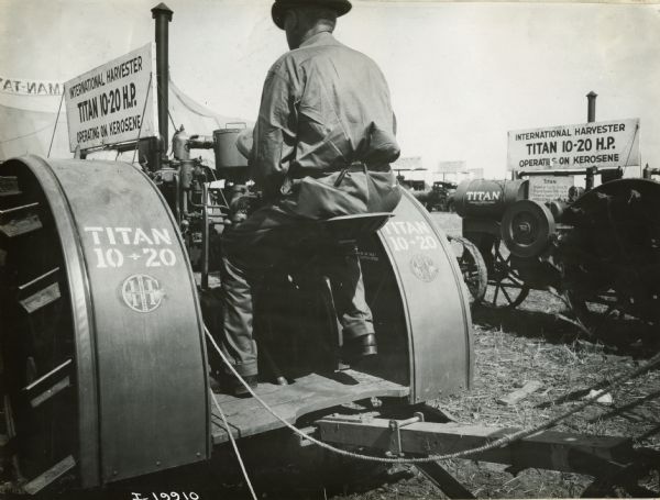 A man is sitting on the Titan 10-20 demonstrating its operation. A sign on top of the tractor reads: "International Harvester Titan 10-20 H.P. Operating on Kerosene." Decals and stenciling are clearly visible on the rear fenders. The demonstration may be part of an agricultural exhibition or fair.