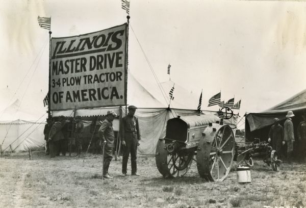 Two men look over a tractor called the Indiana, while behind them a group gathers in front of a large Illinois Master Drive 3-4 Plow Tractor of America tent.