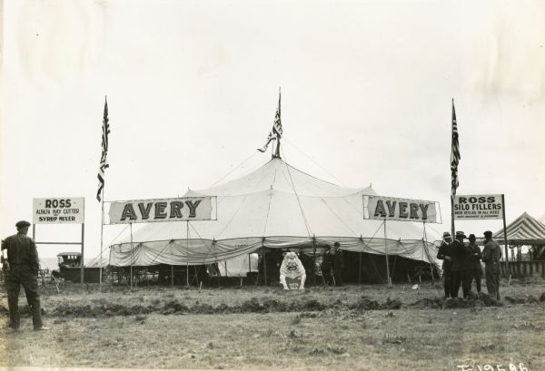A large white tent marked with multiple signs reading: "Avery." Other signs advertise for the "Ross Alfalfa Hay Cutter and Syrup Mixer" and the "Ross Silo Fillers". A large bulldog statue is in the center of the tent holding a sign saying, "Teeth Talk". The tent may have been part of an agricultural exhibit or fair.