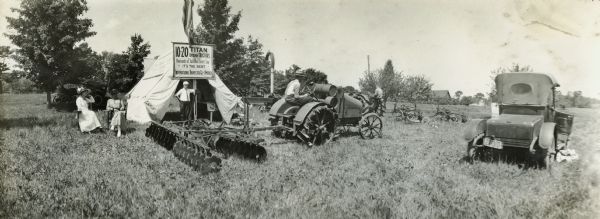A small group of women and men gather around a tent with a sign boasting of satisfied users of the Titan 10-20 Kerosene tractor. Another man is sitting in the seat of what appears to be an International 8-16 tractor with an attached plow.