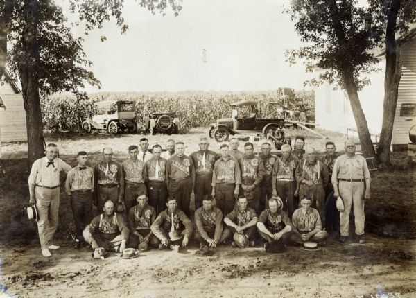 A group of men standing together in front of a farm field all wearing shirts that say "Lincoln Salesman." The men were likely salesmen for the International Harvester Company, employed through the company's Lincoln, Nebraska branch house.