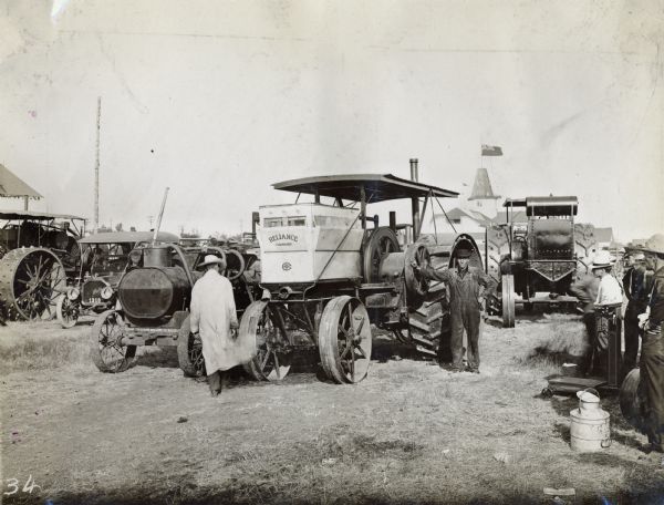 Two men stand near a large Reliance tractor from Milwaukee that is surrounded by many other tractors and vehicles. The tractor was likely part of a tractor demonstration or show attended by various manufacturers (possibly the Fremont Tractor Show at Fremont, Iowa).