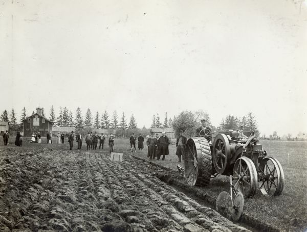A group of men and women gather in a farm field to watch a demonstration of plowing with a Mogul 8-16 tractor.