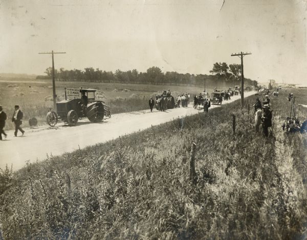 A parade of tractors (including the International Harvester Mogul 12-25), trucks and people moving down a dirt road, possibly as part of a tractor demonstration.