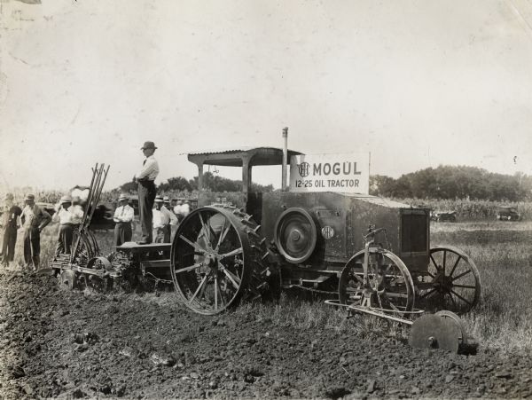 Men stand around an International Harvester Mogul 12-25 tractor in a farm field to watch a plowing demonstration.