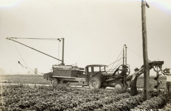 Men working along a road with a modified McCormick-Deering 10-20 industrial tractor with attached trailor and post hole digging rig. The men work for Shippers Rapid Transit and seem to be erecting a telephone or electrical pole.